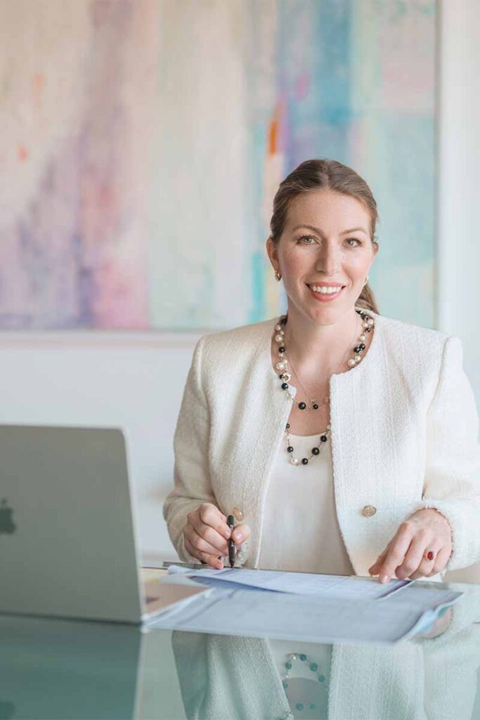 Maja Radisic, founder of Croatia Navigator, smiling warmly at her desk in a white tweed blazer and pearl and black bead necklace, with a MacBook and documents in front of her and a soft pastel abstract painting on the wall behind her.